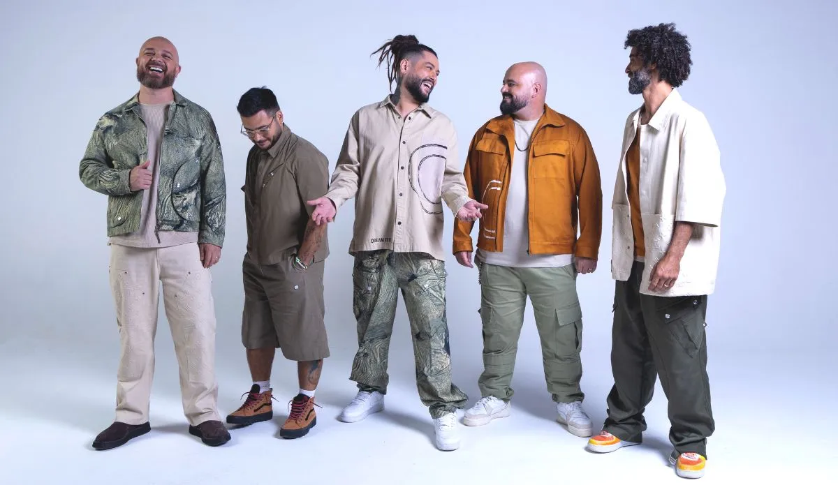 Os cinco integrantes da banda de reggae brasileira Maneva estão alinhados na imagem, vestidos com roupas casuais que refletem diferentes estilos. Eles são Tales de Polli (vocal), Felipe Sousa (guitarra), Fernando Gato (baixo), Fabinho Araújo (bateria) e Diego Andrade (percussão). O fundo da imagem é discreto, destacando os músicos e suas vestimentas.
