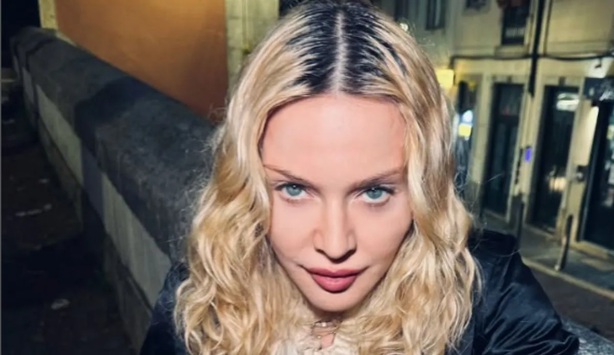 Madonna posa de frente para a câmera em uma rua à noite, com cabelos loiros ondulados e expressão séria.