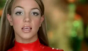 Britney Spears no icônico macacão vermelho no clipe de "Oops!... I Did It Again", com maquiagem dos anos 2000 e fundo desfocado em tons verdes e dourados.