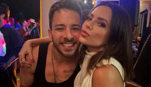 Junior Lima e Sandy posam sorridentes em festa, com luzes coloridas ao fundo e outras pessoas presentes no ambiente