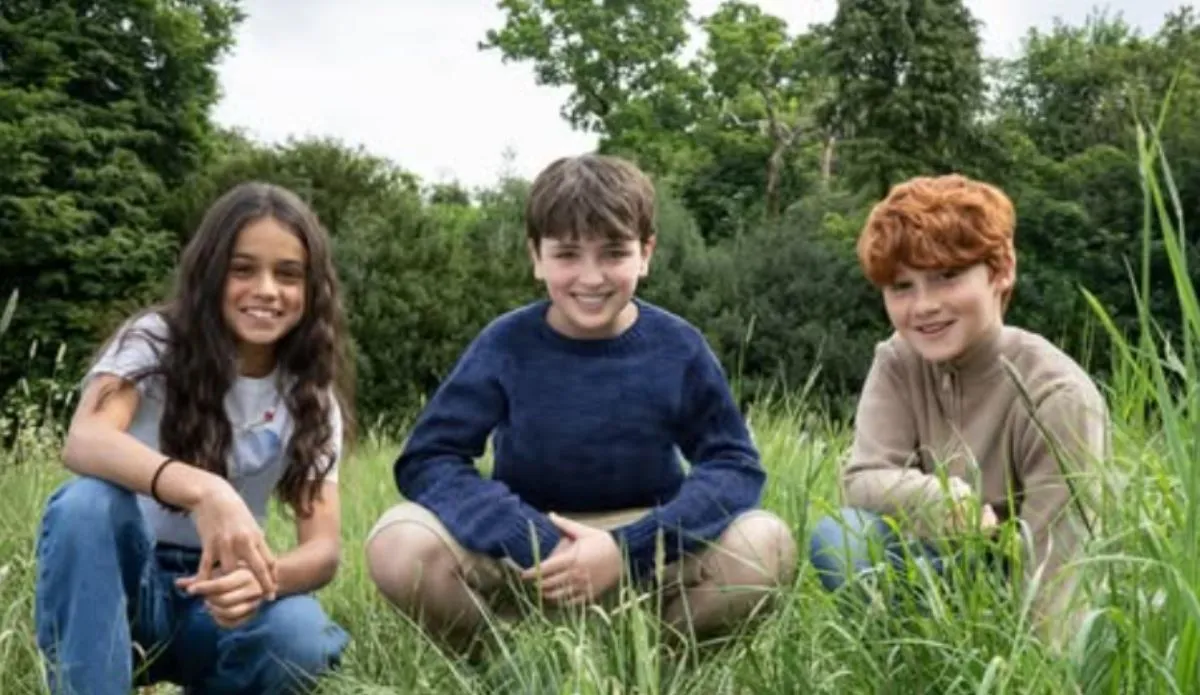 Trio de jovens atores da nova série de Harry Potter posa sorridente sentado na grama, com árvores ao fundo — Arabella Stanton (Hermione), Dominic McLaughlin (Harry) e Alastair Stout (Ron).