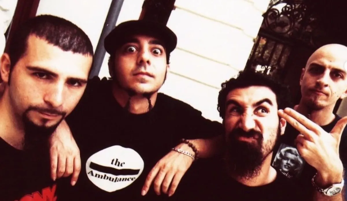 Imagem dos integrantes da banda System of a Down. O vocalista Serj Tankian, o guitarrista Daron Malakian, o baixista Shavo Odadjian e o baterista John Dolmayan posam juntos em frente a uma parede branca e um portão de ferro