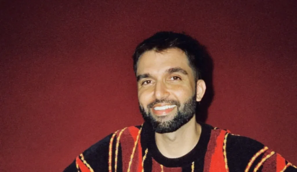 Silva, nova atração confirmada no Coala Festival 2025, sorri em frente a uma parede vermelha, vestindo uma blusa colorida listrada em tons de vermelho, amarelo e preto.