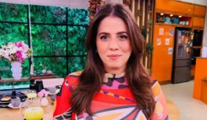 Tati Machado em um estúdio de programa de TV, usando uma blusa colorida com estampa abstrata, cabelos soltos e maquiagem suave. Ao fundo, decoração com flores, mesa com bebidas e cozinha com tons de laranja.