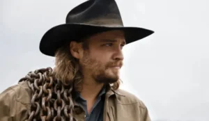 Luke Grimes caracterizado como Kayce Dutton em cena de Yellowstone, usando chapéu de cowboy e carregando uma corrente de ferro no ombro