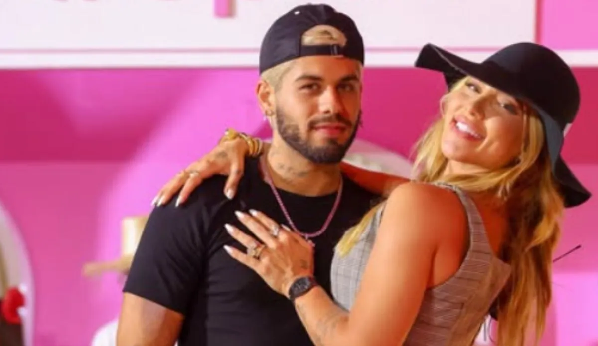 Zé Felipe e Virgínia Fonseca posam juntos sorrindo, em evento com fundo rosa; ela usa chapéu preto e vestido cinza, ele veste camiseta preta e boné.