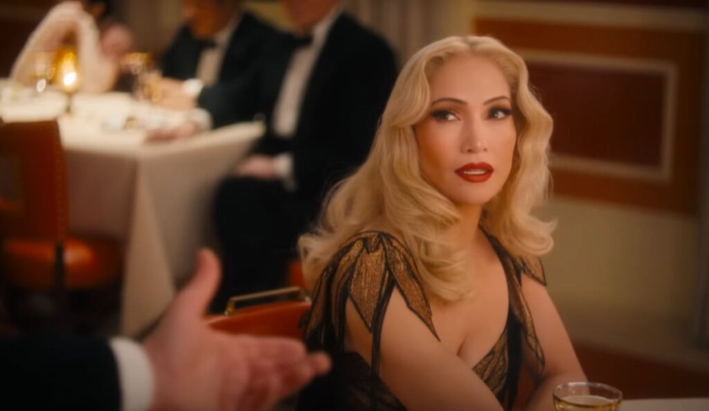 Jennifer Lopez caracterizada como Ingrid Luna, em cena do remake musical de “O Beijo da Mulher-Aranha”, vestindo um glamouroso vestido dourado com detalhes pretos e maquiagem marcante, em um ambiente de jantar sofisticado.