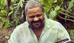 Arlindo Cruz sorri enquanto olha para baixo. Ele veste uma camisa verde clara estampada e está em um ambiente externo com vegetação ao fundo.