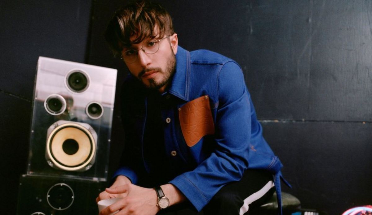 bbno$, rapper canadense, posa sentado ao lado de uma caixa de som grande; ele veste jaqueta azul com detalhe marrom, óculos redondos e tem cabelo castanho com franja.