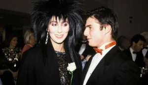 Cher e Tom Cruise durante um evento nos anos 1980, lado a lado e sorrindo; Cher usa um vestido preto brilhante e penteado volumoso, enquanto Tom aparece de smoking com gravata borboleta vermelha.