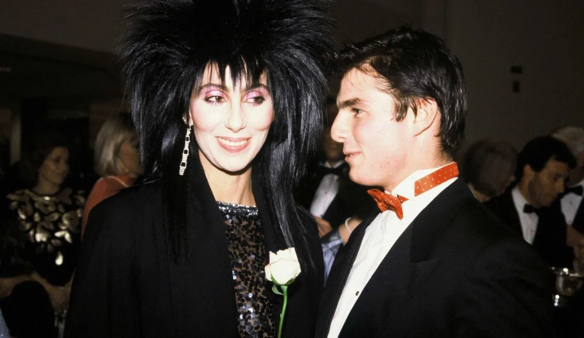 Cher e Tom Cruise durante um evento nos anos 1980, lado a lado e sorrindo; Cher usa um vestido preto brilhante e penteado volumoso, enquanto Tom aparece de smoking com gravata borboleta vermelha.