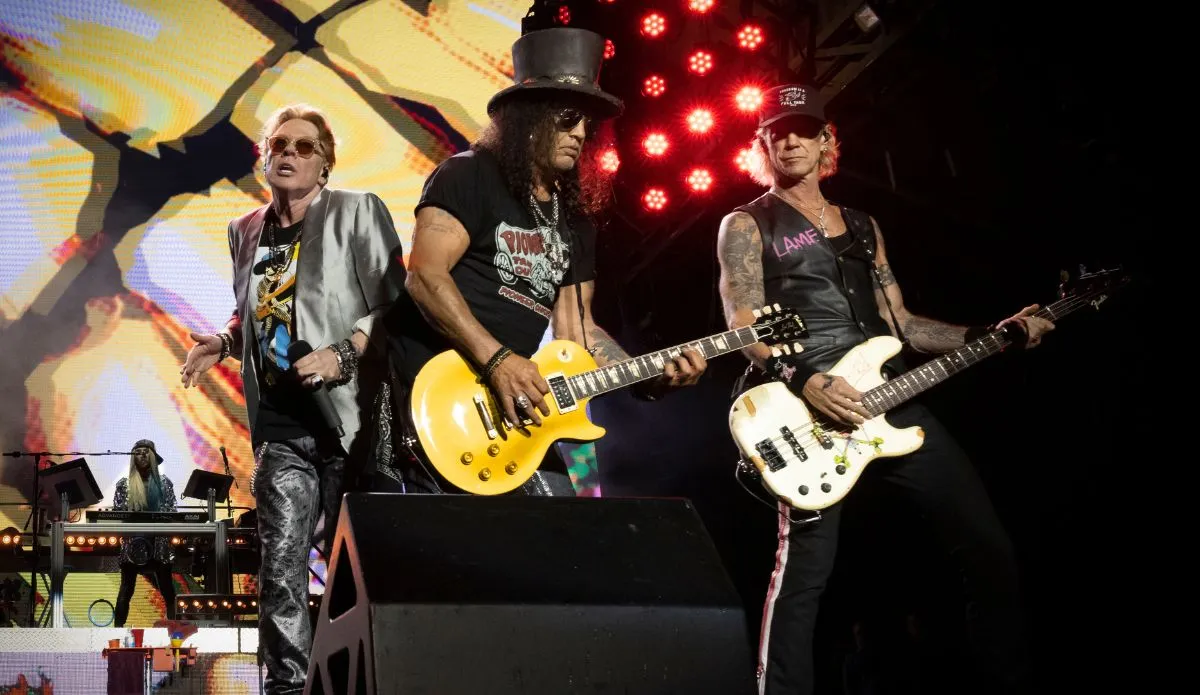Integrantes da banda Guns N’ Roses durante apresentação ao vivo: Axl Rose (vocais), Slash (guitarra) e Duff McKagan (baixo) se apresentam em palco com telão colorido e iluminação intensa.