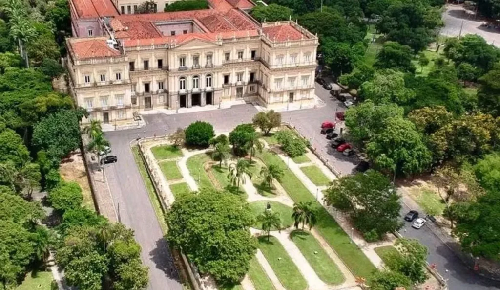 Vista aérea do prédio histórico do Museu Nacional, localizado na Quinta da Boa Vista, no Rio de Janeiro. O edifício em estilo neoclássico é cercado por jardins simétricos, com caminhos de terra, palmeiras e áreas verdes. Ao redor, há árvores e algumas vagas de estacionamento com carros estacionados.