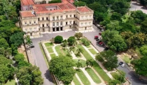 Vista aérea do prédio histórico do Museu Nacional, localizado na Quinta da Boa Vista, no Rio de Janeiro. O edifício em estilo neoclássico é cercado por jardins simétricos, com caminhos de terra, palmeiras e áreas verdes. Ao redor, há árvores e algumas vagas de estacionamento com carros estacionados.
