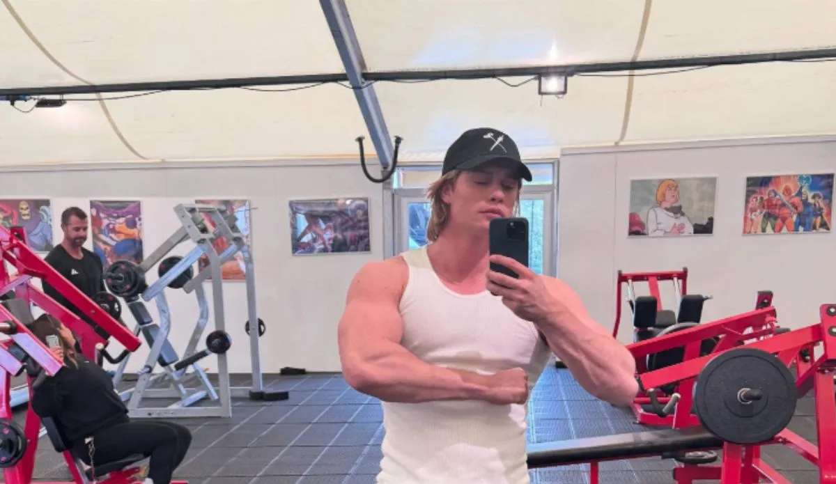Nicholas Galitzine tira selfie em frente ao espelho da academia, vestindo regata branca e boné preto, mostrando os músculos durante preparação física para viver He-Man. Ao fundo, pôsteres de personagens de "Mestres do Universo" decoram a parede.