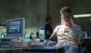 Cena do trailer da série The Institute (MGM+, inspirada no livro de Stephen King), mostrando o ator Joe Freeman como o adolescente Luke Ellis. Ele está sentado em uma cadeira médica, com sensores no braço e no peito, conectado a um monitor de sinais vitais, em um ambiente clínico que sugere a instalação do Instituto, onde crianças com poderes telepáticos e telecinéticos são mantidas.