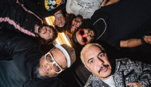 Integrantes do Planet Hemp posam reunidos em círculo, olhando para a câmera de cima para baixo; na frente, Marcelo D2 aparece com cabelo descolorido e jaqueta estampada, ao lado de BNegão, que usa boné e óculos brancos. Ao fundo, outros membros completam a formação com expressões sérias e estilo marcante.
