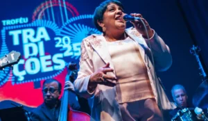 Rosa Passos canta ao microfone durante apresentação na cerimônia do Troféu Tradições UBC 2025, vestindo um conjunto claro com detalhes bordados; ao fundo, músicos a acompanham no palco e o logotipo do evento aparece iluminado.