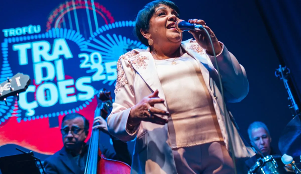 Rosa Passos canta ao microfone durante apresentação na cerimônia do Troféu Tradições UBC 2025, vestindo um conjunto claro com detalhes bordados; ao fundo, músicos a acompanham no palco e o logotipo do evento aparece iluminado.