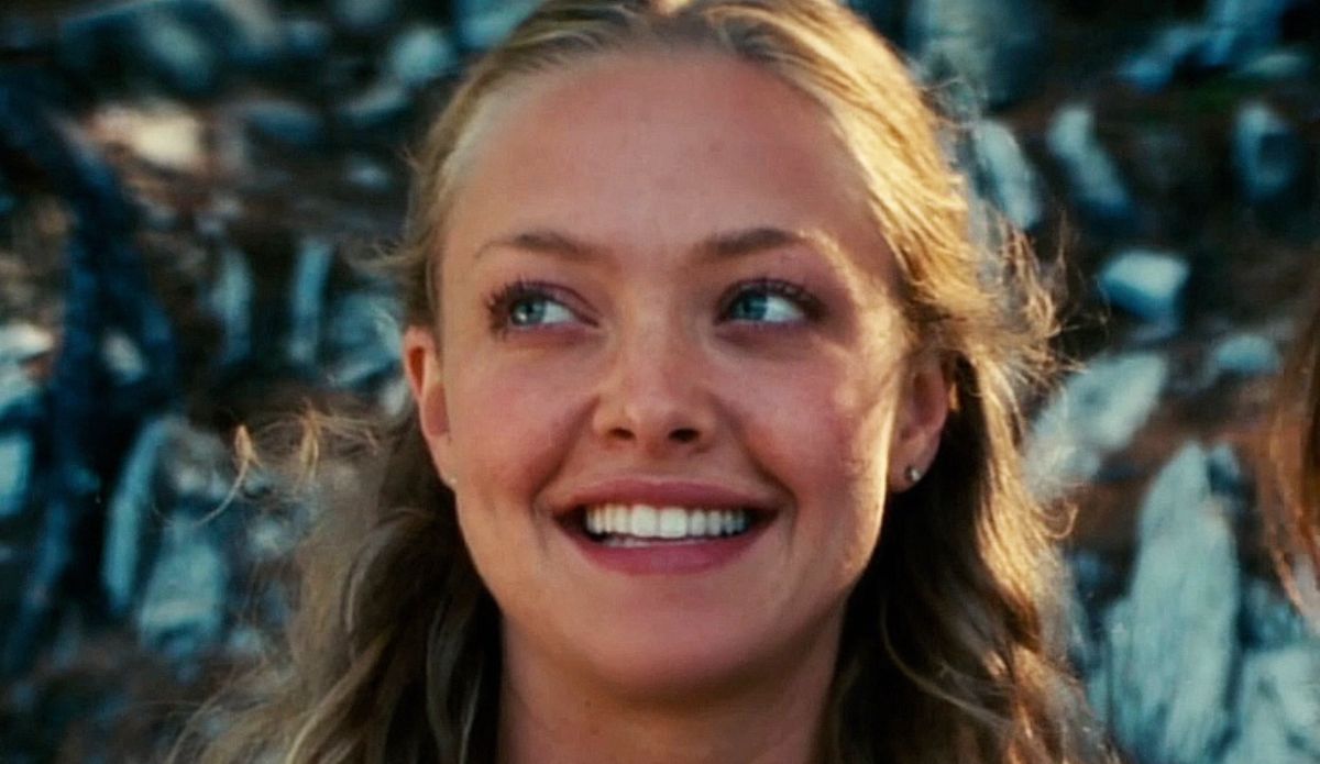 Amanda Seyfried sorrindo em cena do filme Mamma Mia!, com cabelos ondulados e loiros ao vento, em cenário ao ar livre com pedras ao fundo.