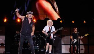 Integrantes do AC/DC se apresentam em palco iluminado, com Brian Johnson cantando ao microfone, Angus Young tocando guitarra com seu icônico uniforme escolar e outros músicos ao fundo; telão exibe close vibrante da performance ao vivo.