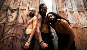 Integrantes da banda Black Pantera posam sem camisa em frente a uma porta de madeira envelhecida com grafites; todos encaram a câmera com expressão séria e postura firme.