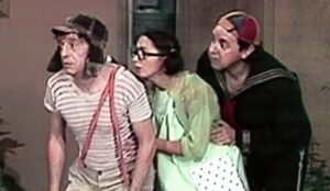 Alt text: Chaves, Chiquinha e Quico observam algo com expressão curiosa em cena do seriado, vestindo seus trajes clássicos no pátio da vila.