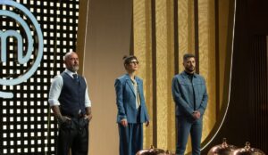 Jurados do MasterChef Brasil 2025 em pé no estúdio do programa. Da esquerda para a direita, um Henrique Fogaça com barba grisalha veste colete e camisa branca; Helena Rizzo usa um coque alto, batom vermelho e óculos escuros usa terno azul com gravata amarela; e Evaristo Costa de barba curta veste conjunto jeans azul. Ao fundo, o logo do MasterChef e painéis dourados iluminam o cenário.
