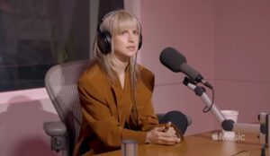 Hayley Williams durante entrevista no estúdio da Apple Music. A vocalista do Paramore aparece sentada, usando fones de ouvido e blazer marrom, diante de um microfone profissional.