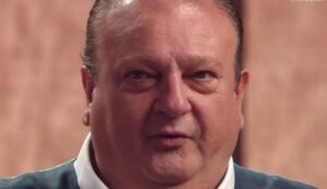 Close de Érick Jacquin no estúdio do MasterChef Brasil. O chef francês aparece com expressão séria, vestindo camisa branca e suéter azul-escuro, com o fundo desfocado em tom amadeirado.