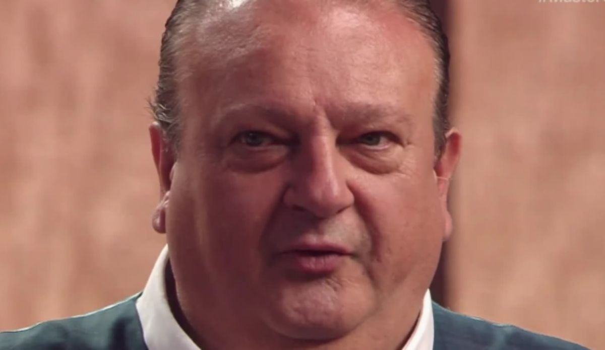 Close de Érick Jacquin no estúdio do MasterChef Brasil. O chef francês aparece com expressão séria, vestindo camisa branca e suéter azul-escuro, com o fundo desfocado em tom amadeirado.