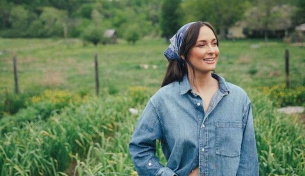 Kacey Musgraves sorri em meio a um campo verde, vestindo uma camisa jeans e lenço azul nos cabelos. Ao fundo, vegetação e árvores completam o cenário rural.