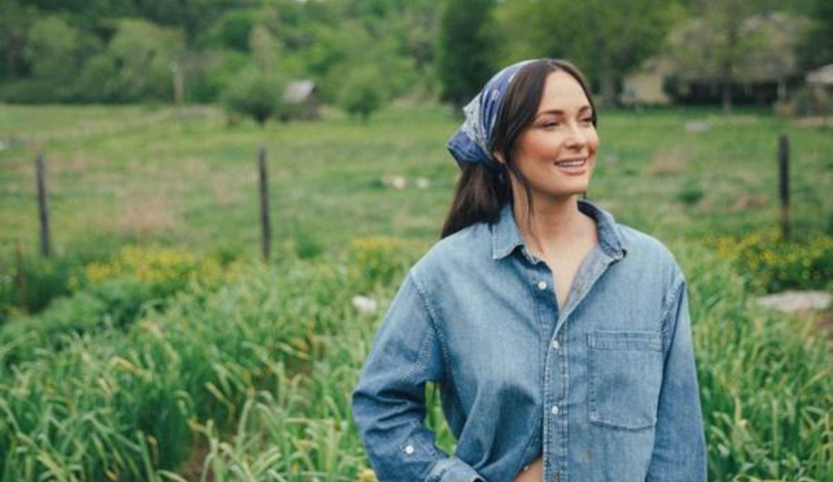 Kacey Musgraves sorri em meio a um campo verde, vestindo uma camisa jeans e lenço azul nos cabelos. Ao fundo, vegetação e árvores completam o cenário rural.
