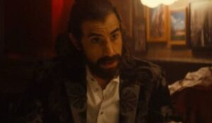 Mephisto, personagem do MCU interpretado por Sacha Baron Cohen, aparece com cabelos longos e barba escura, vestindo paletó estampado e camisa branca, sentado em ambiente com iluminação quente e quadros ao fundo.