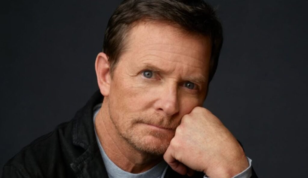 Michael J. Fox em foto promocional com expressão séria, apoiando o rosto na mão, em frente a um fundo escuro.