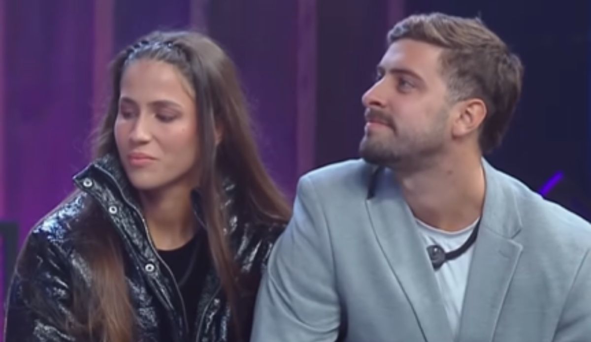 Natália e Eike sentados lado a lado durante programa ao vivo do Power Couple Brasil 7. Natália veste uma jaqueta preta brilhante e olha discretamente para o lado. Eike, com blazer cinza claro, observa Natália com expressão serena.