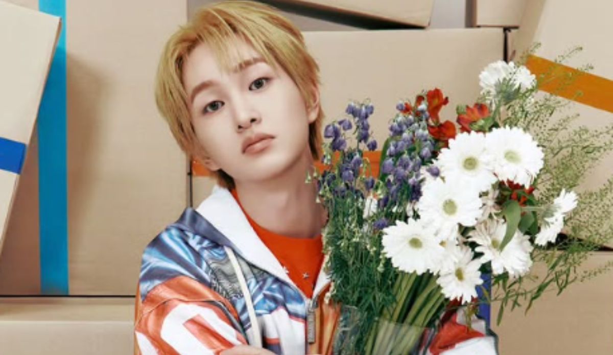 Onew, integrante do SHINee, posa segurando um buquê de flores com margaridas brancas, lavandas e flores vermelhas. Ele veste uma jaqueta colorida e olha diretamente para a câmera, com expressão suave. Ao fundo, há várias caixas de papelão empilhadas.