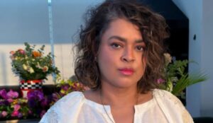 Preta Gil em close, usando uma blusa branca com maquiagem leve e expressão serena, em frente a um arranjo de flores coloridas. A luz do fim de tarde ilumina seu rosto.