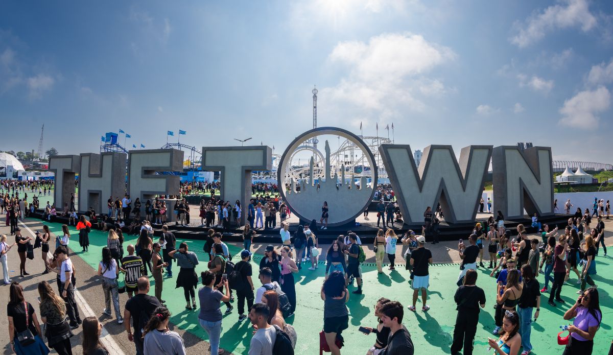 Multidão circula e tira fotos em frente à instalação com letras gigantes que formam a palavra "The Town", no centro da Cidade da Música, durante o festival em São Paulo. O dia está ensolarado, com céu azul e poucas nuvens. Ao fundo, estruturas de brinquedos e bandeiras azuis compõem o cenário.