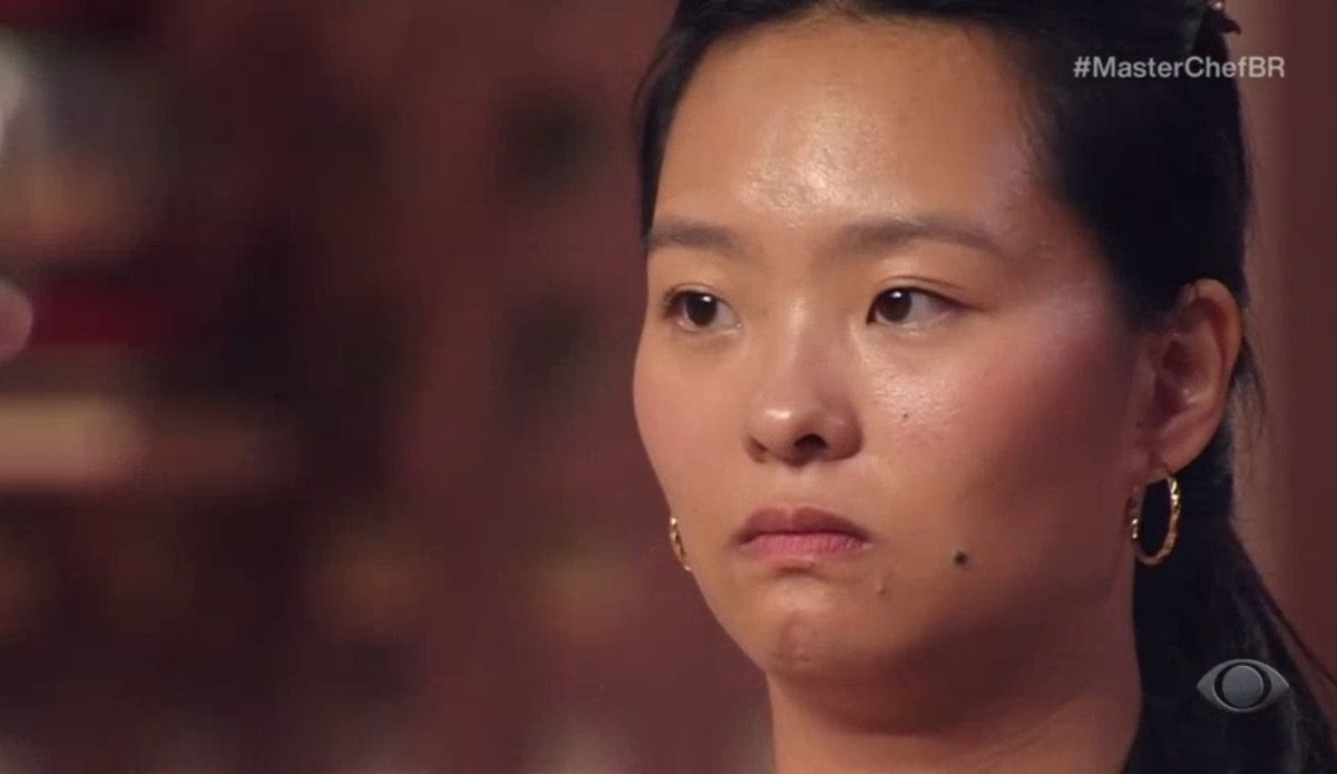 Teresa, participante do MasterChef Brasil 2025, observa séria e concentrada durante o anúncio do resultado da repescagem. Ela usa brincos dourados e está com expressão tensa, em destaque no estúdio do programa.