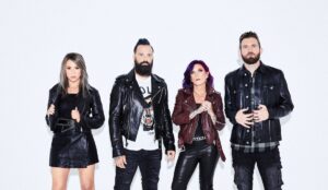 Integrantes da banda Skillet posam em frente a um fundo branco. Da esquerda para a direita: Jen Ledger veste jaqueta e saia preta de couro; John Cooper usa camiseta estampada e jaqueta preta; Korey Cooper aparece com cabelo roxo, jaqueta vinho e calça de couro; e Seth Morrison veste jaqueta preta sobre camisa xadrez escura. Todos encaram a câmera com expressão séria.