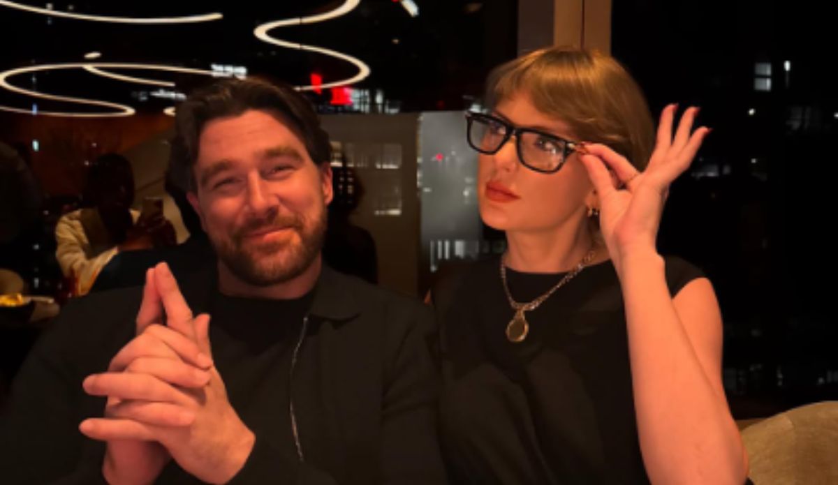 Taylor Swift e Travis Kelce entadas lado a lado em um ambiente fechado e elegante, possivelmente um restaurante.À direita, Swift, uma mulher loira - com franja , batom vermelho, óculos de armação grossa e roupa preta — segura os óculos com uma expressão levemente irônica ou analítica. À esquerda, Kelce, de barba curta e cabelo penteado para o lado sorri discretamente, com as mãos juntas. O fundo tem iluminação baixa e moderna, com luzes em espiral no teto e reflexos de janelas que sugerem uma cidade à noite.