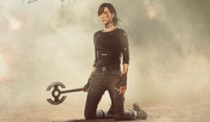 Imagem mostra a personagem Andy (Charlize Theron) ajoelhada no chão em meio à fumaça e explosões, com o corpo coberto de ferimentos e sujeira. Ela veste roupas pretas rasgadas e segura seu machado duplo, símbolo de sua trajetória como imortal em "The Old Guard 2".