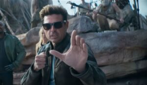 Karl Urban como Johnny Cage em "Mortal Kombat 2", usando óculos escuros e jaqueta de couro, em pose de luta durante uma cena no deserto, com personagens armados ao fundo.