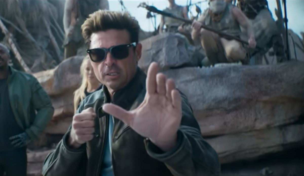 Karl Urban como Johnny Cage em "Mortal Kombat 2", usando óculos escuros e jaqueta de couro, em pose de luta durante uma cena no deserto, com personagens armados ao fundo.