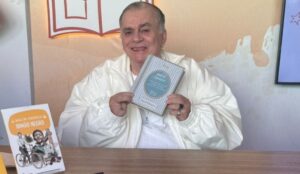 Walcyr Carrasco sorri enquanto segura um de seus livros durante evento literário. Ele veste uma jaqueta branca e está sentado à mesa, com outro livro seu, intitulado "Irmão Negro", exposto ao lado.