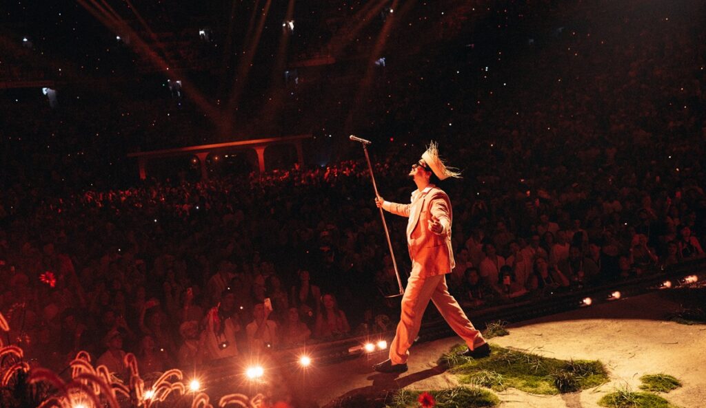 Foto tirada do Bad Bunny, ao fundo do palco de sua residência em Porto Rico. Benito está de lado, olhando para esquerda, segurando o suporte de microfone e ao fundo, o publico.