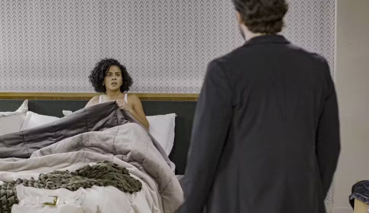 Maria de Fátima (Bella Campos) entada na cama, assustada, segurando um cobertor, enquanto Afonso (Humberto Carrão) aparece de costas, usando terno, está em pé diante dela no quarto em cena de Vale Tudo