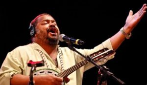 Arlindo Cruz cantando ao vivo com expressão intensa, usando camisa clara, fones de ouvido e tocando cavaquinho diante de um microfone, com fundo preto.