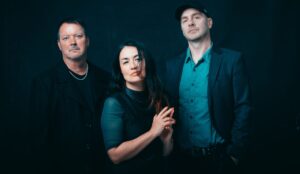 Integrantes da banda britânica Bôa posam juntos em fundo escuro. Jasmine Rodgers está ao centro, com expressão séria e mãos entrelaçadas; à esquerda, Alex Caird veste roupa preta; à direita, Lee Sullivan usa boné, camisa azul e blazer listrado.